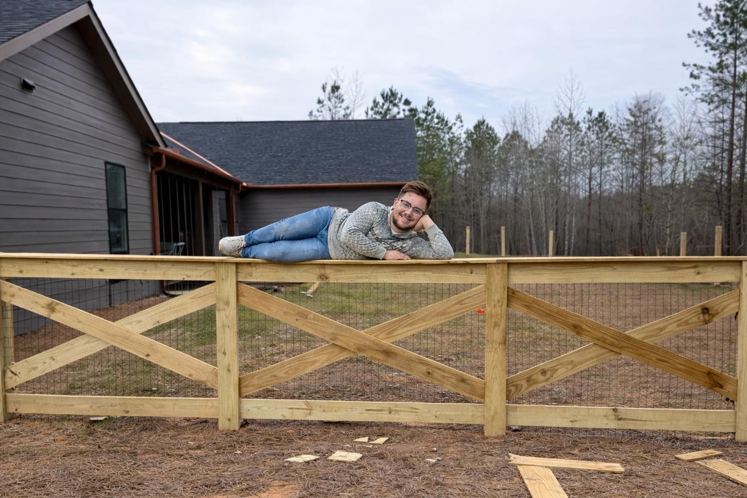 A person wearing a grey sweater and blue jeans lies resting on their side along the top rail of a wooden fence.
