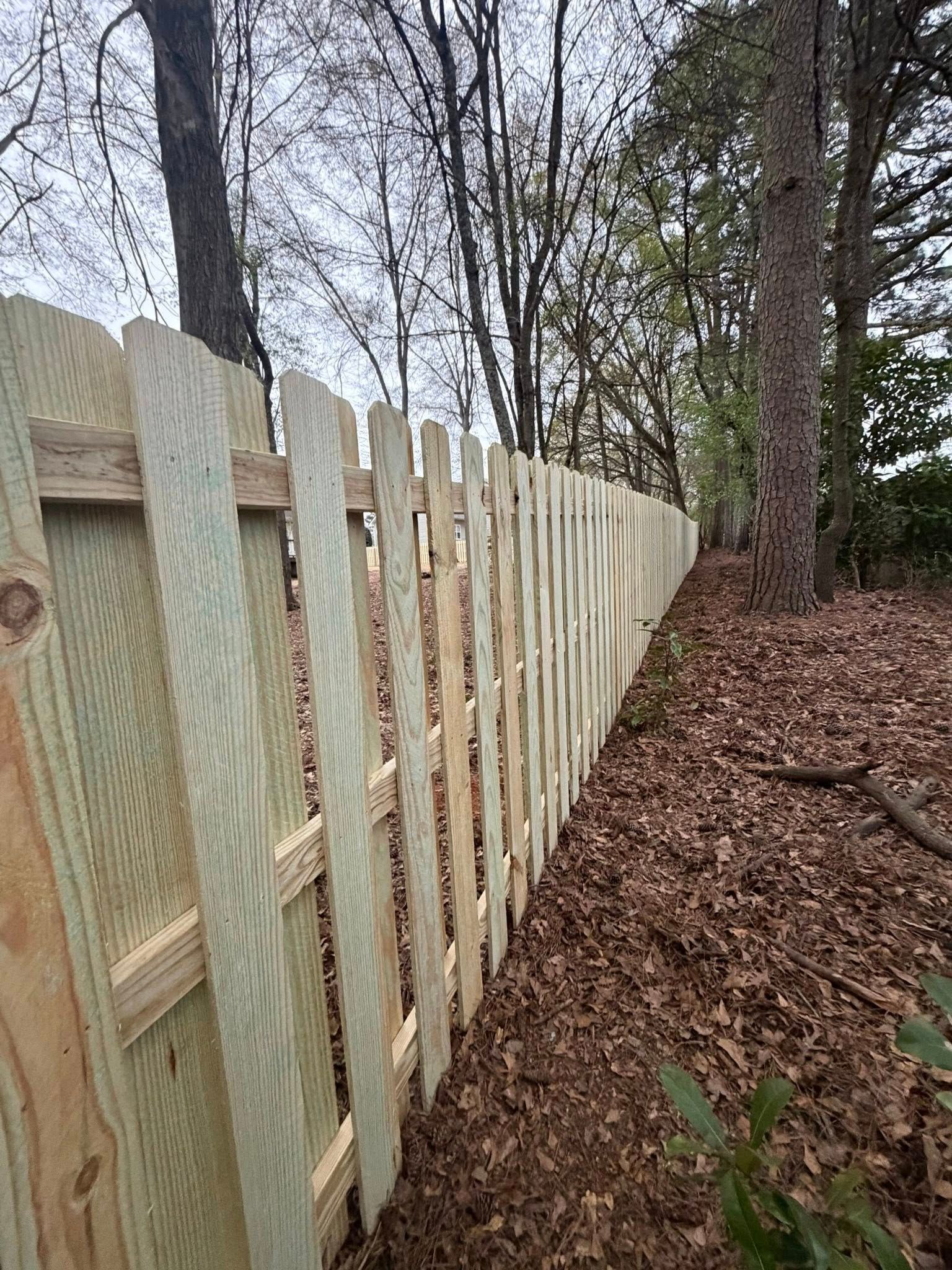 A perspective view of a newly installed wooden picket fence running along a wooded area covered in fallen brown leaves.
