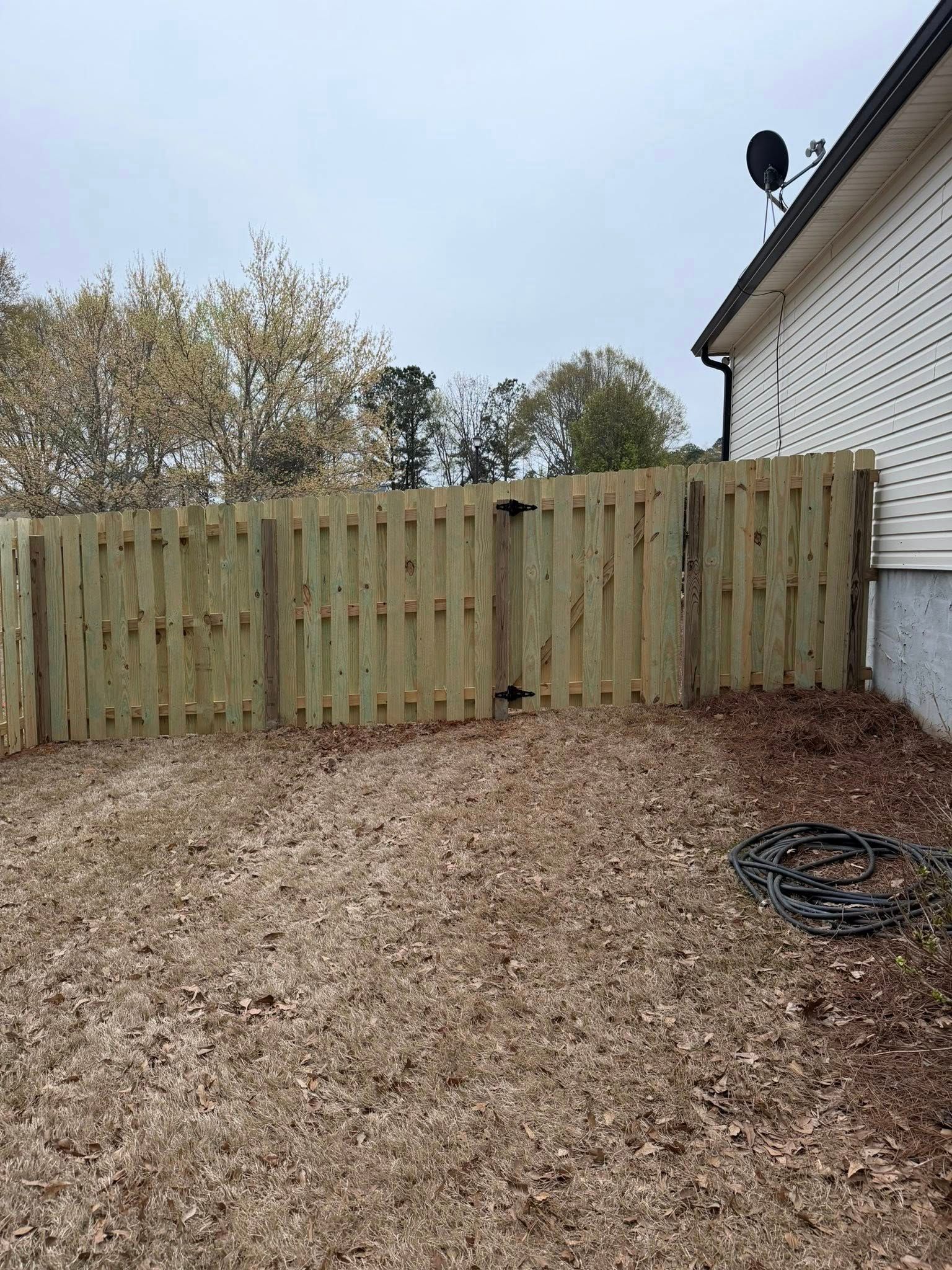 A wooden privacy fence stands in a backyard covered in fallen leaves, adjacent to the white siding of a house.