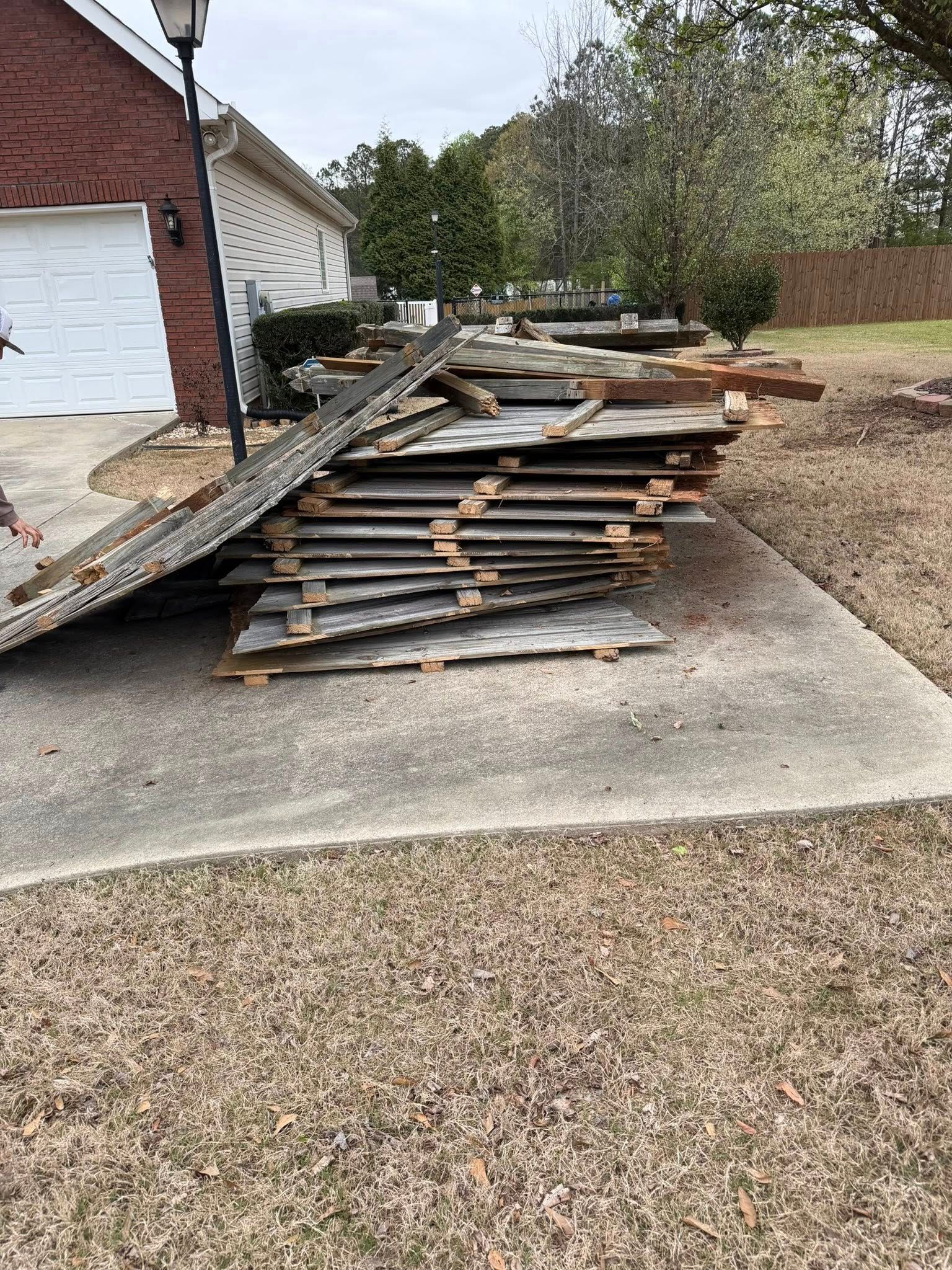 A stack of weathered wooden fence panels sits on a concrete driveway in front of a brick house.