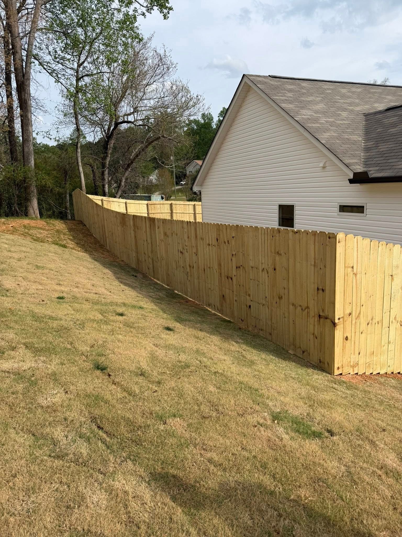 A newly installed light-wood privacy fence runs along a sloped, grassy lawn next to the side of a white house.