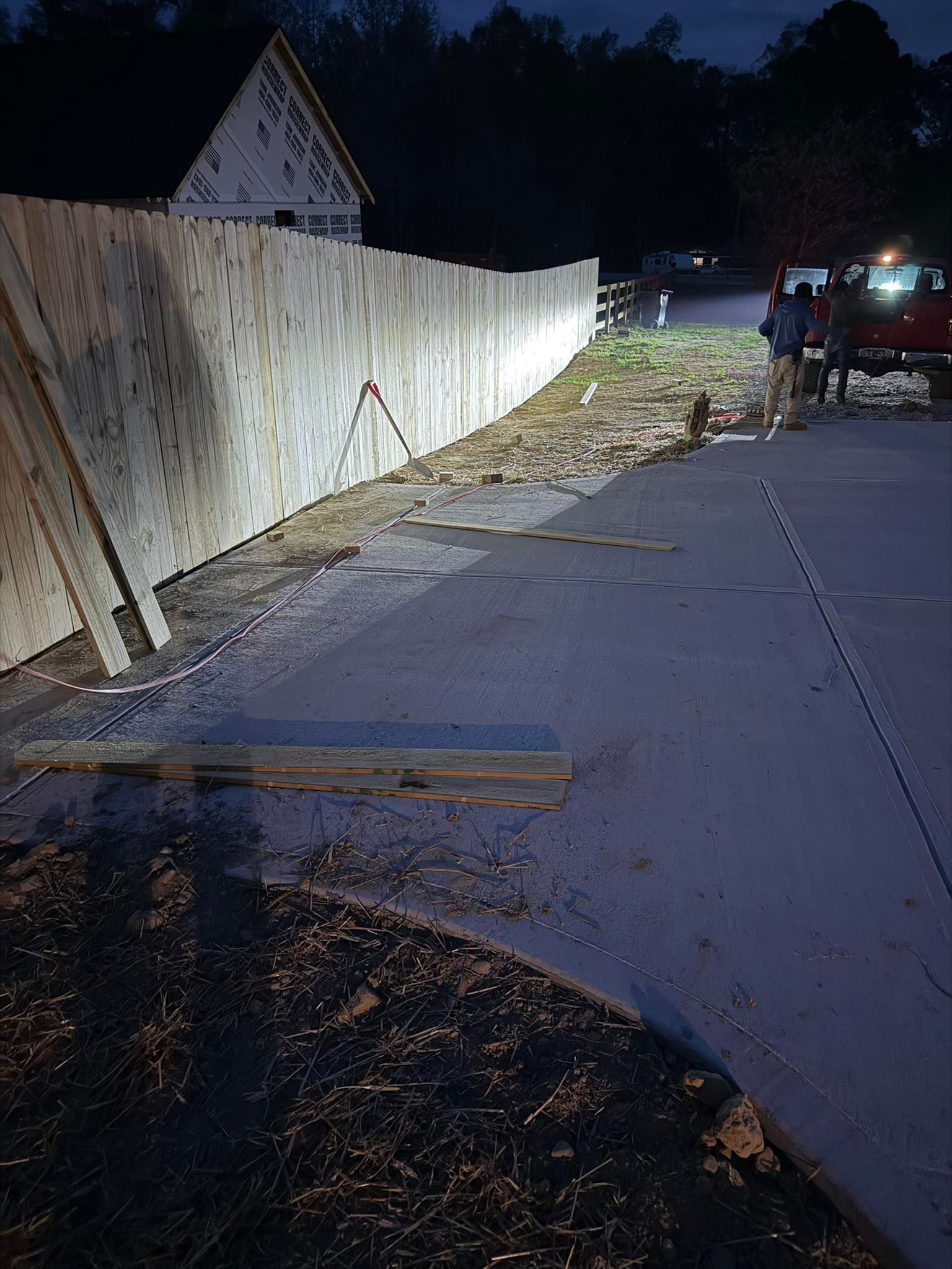 A wooden construction fence lines a dark, outdoor site next to a paved area, with a vehicle visible in the distance.