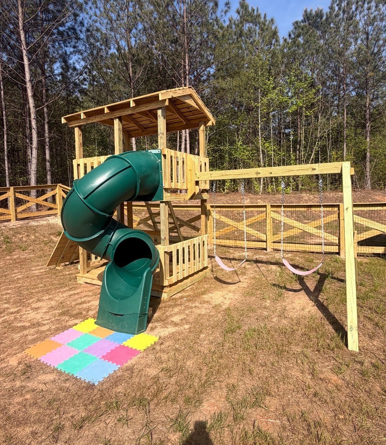 Wooden playground tower with green spiral slide and colorful mat on grassy hillside, trees in background