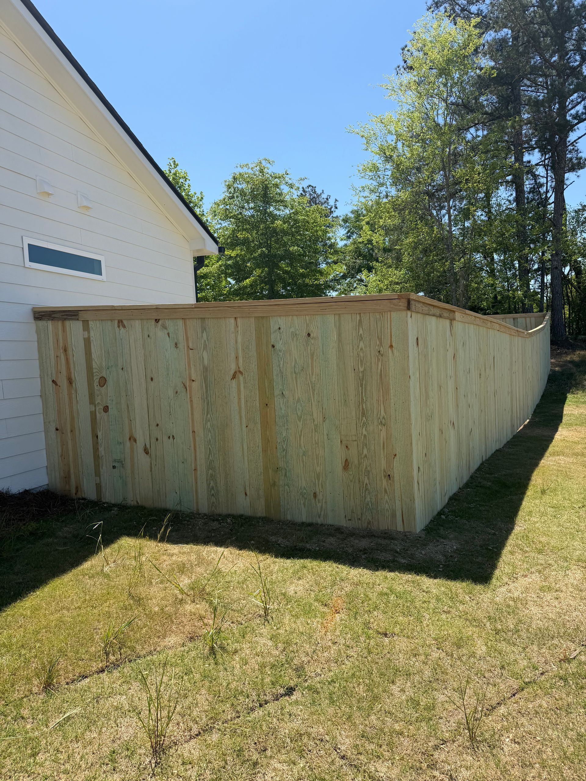 Wooden backyard fence corner beside a white house, with trees and grass in sunlight.