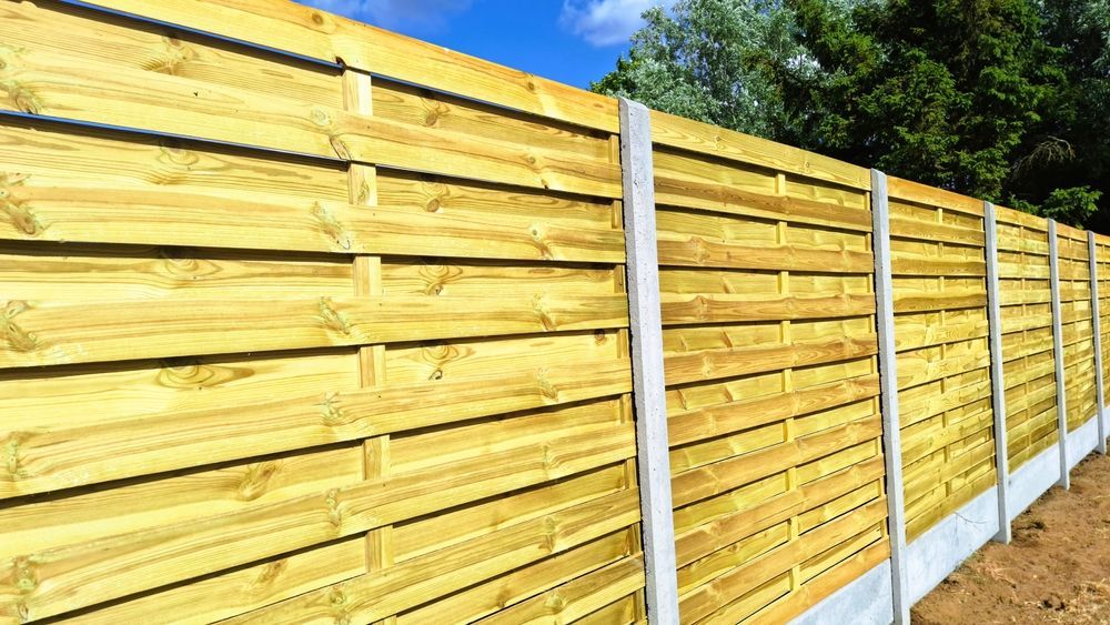A long, horizontal wooden fence with slats, supported by concrete posts and gravel boards against a clear blue sky.