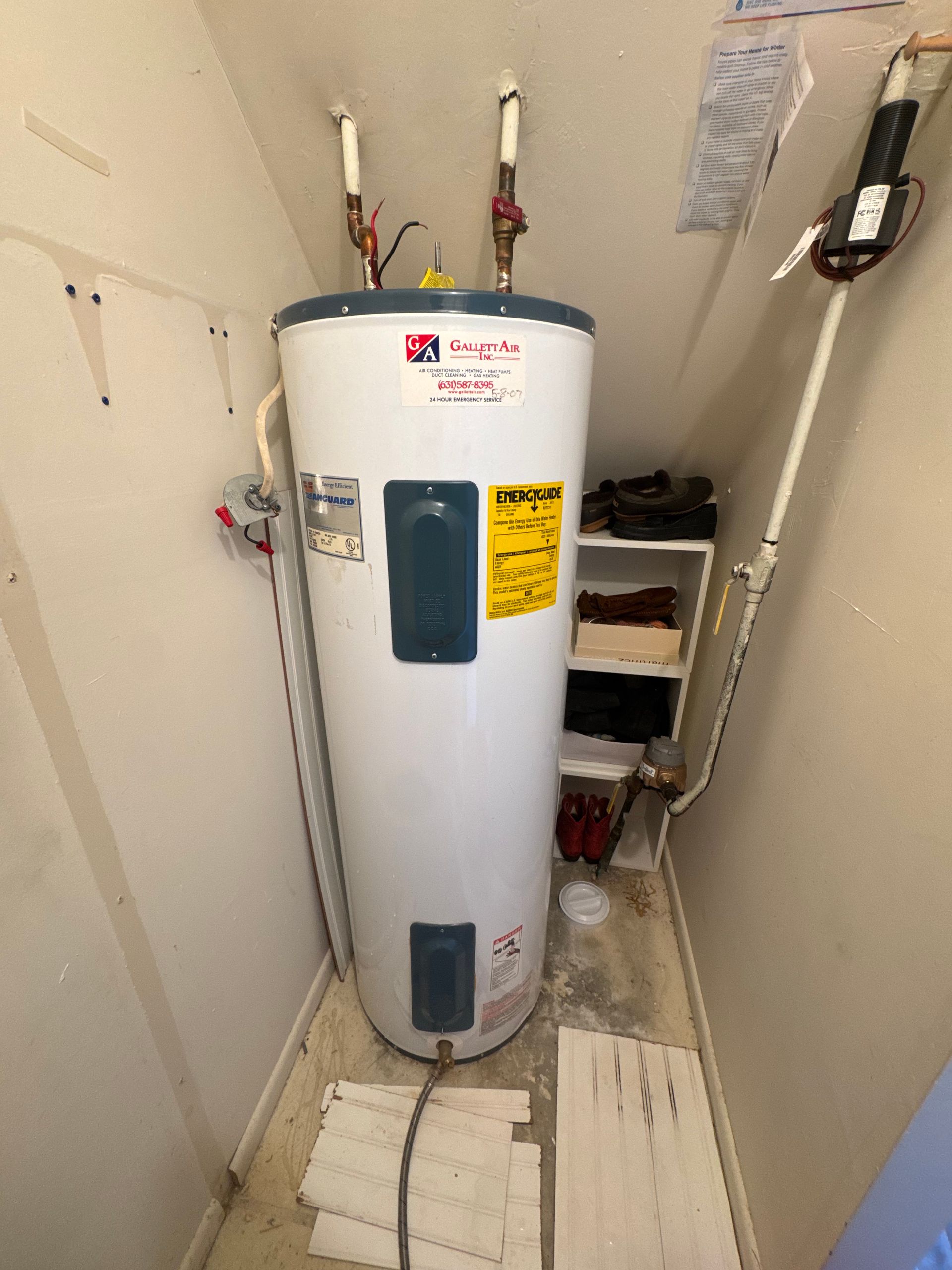 Water heater in a small, enclosed space, with pipes, valves, and a small shelf.