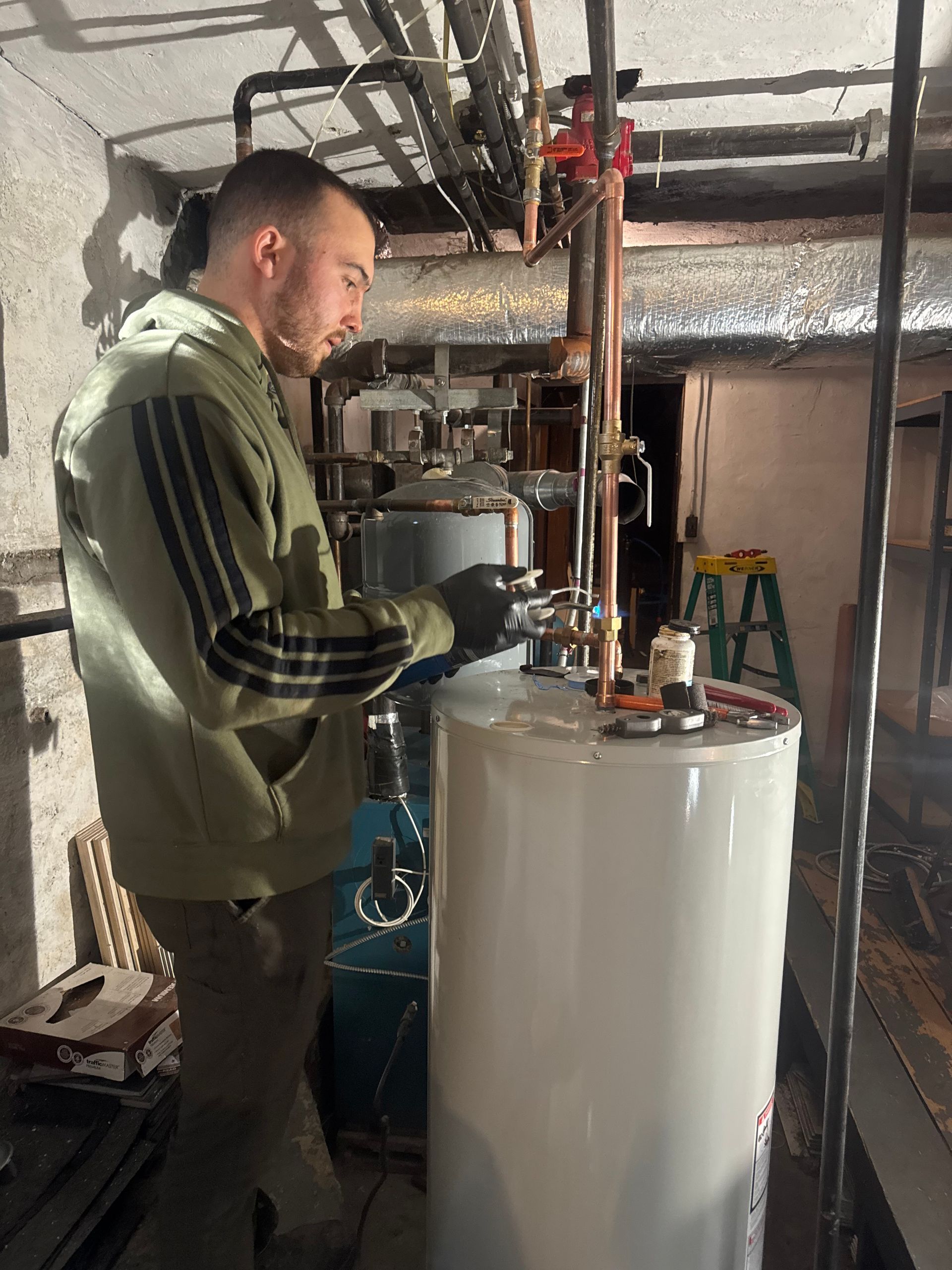 A person in a green jacket works on a hot water tank in a basement.