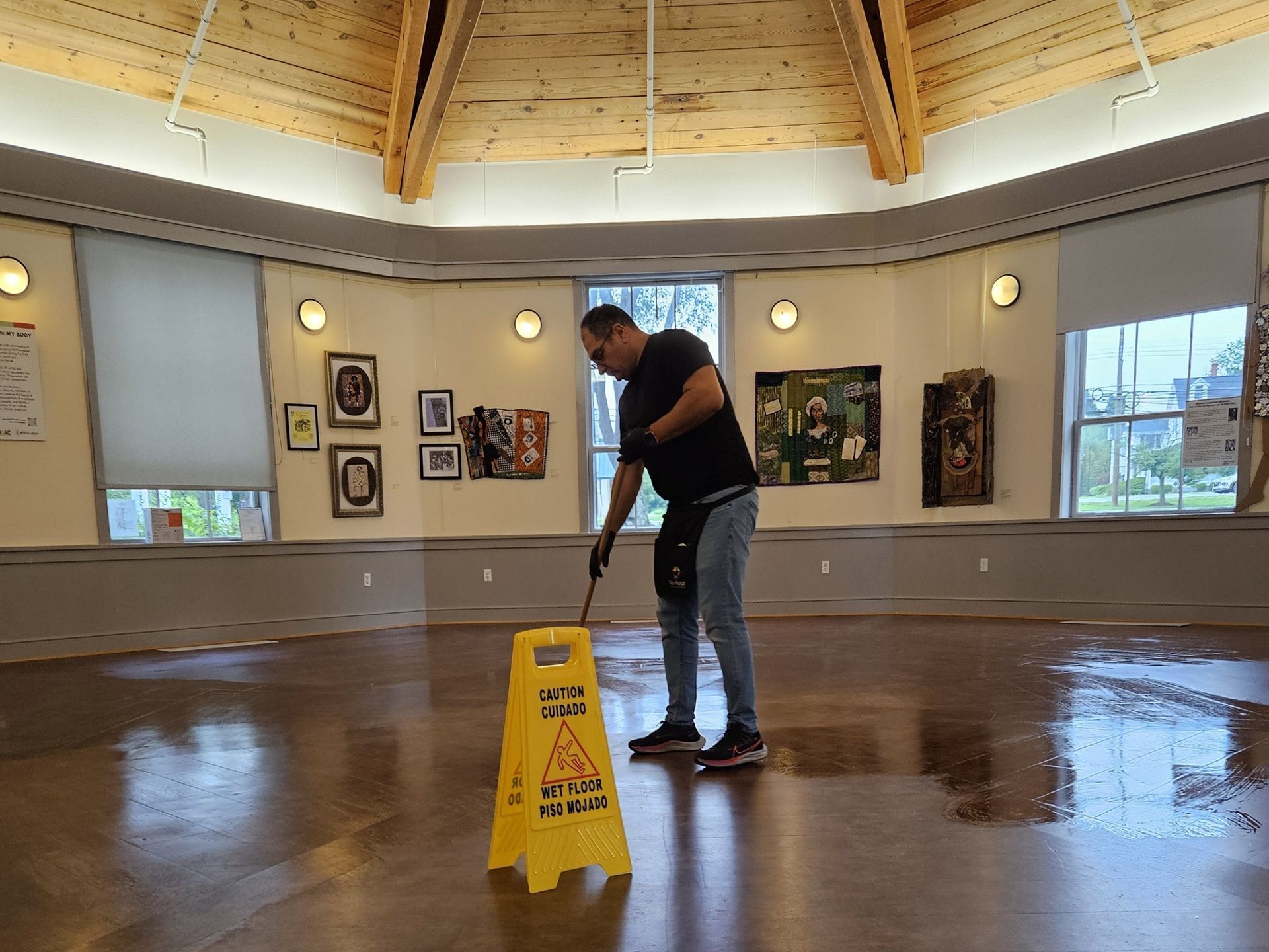 Person mopping a wet floor in a gallery with a caution sign. Wooden floor reflects light. Person mopping a wet floor in a gallery with a caution sign. Wooden floor reflects light.