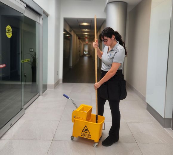 Person mopping a wet floor in a gallery with a caution sign. Wooden floor reflects light.