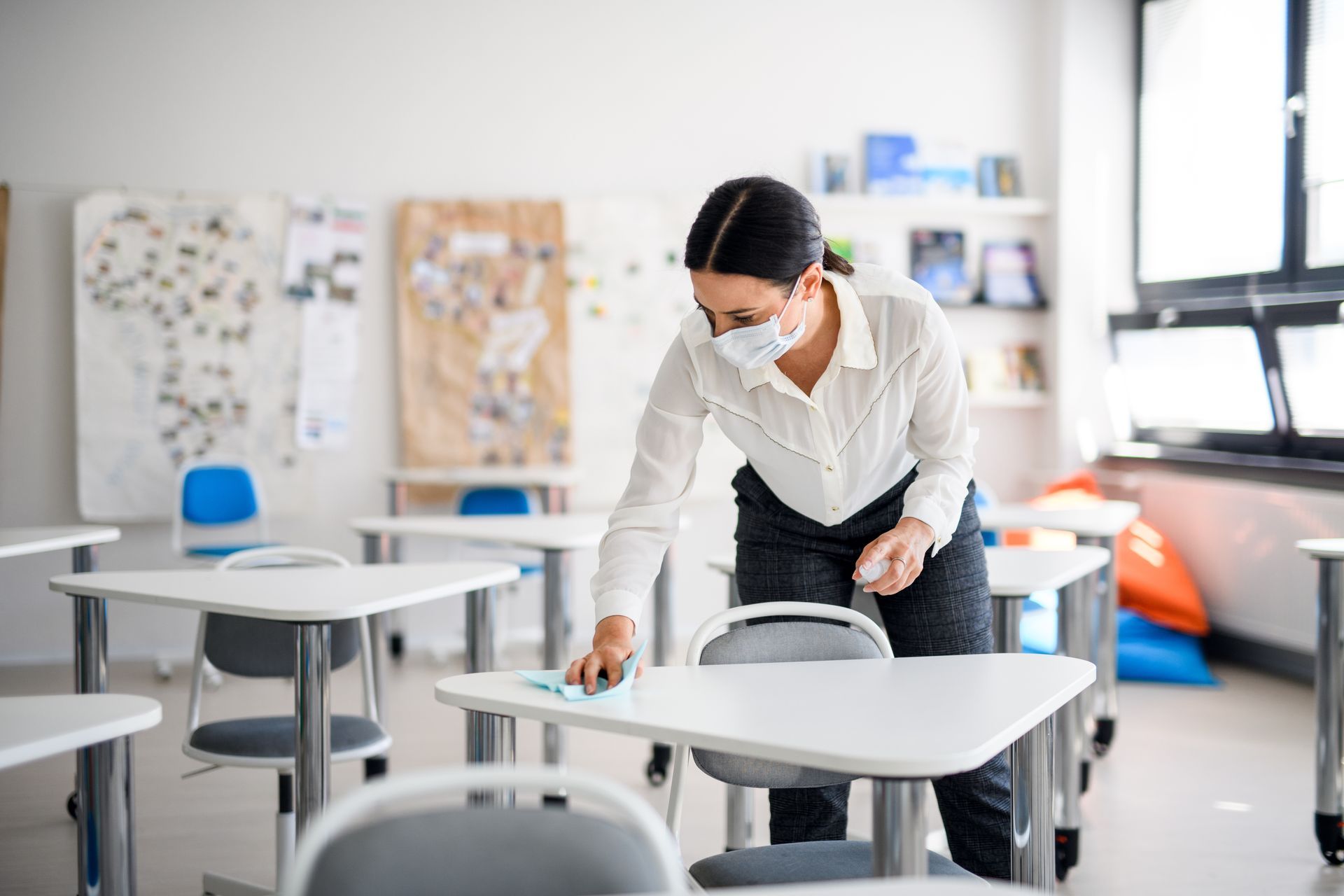 Woman in mask cleaning a desk in a classroom. Woman in mask cleaning a desk in a classroom.