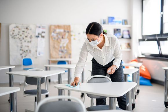 Woman in mask cleaning a desk in a classroom.