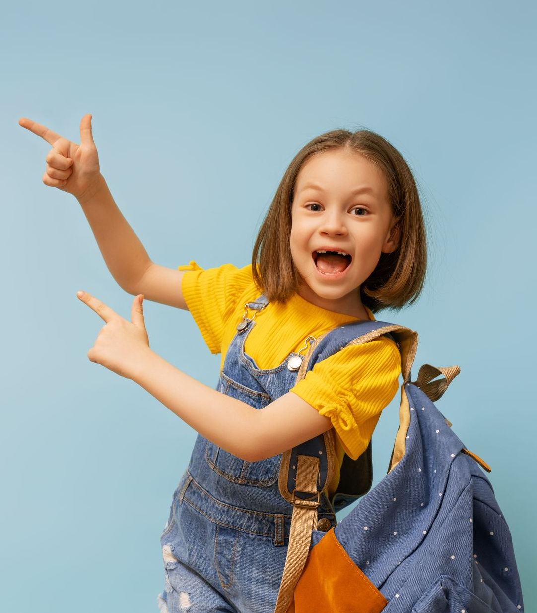 Girl with backpack points and smiles, wearing yellow shirt and blue overalls, against blue backdrop.