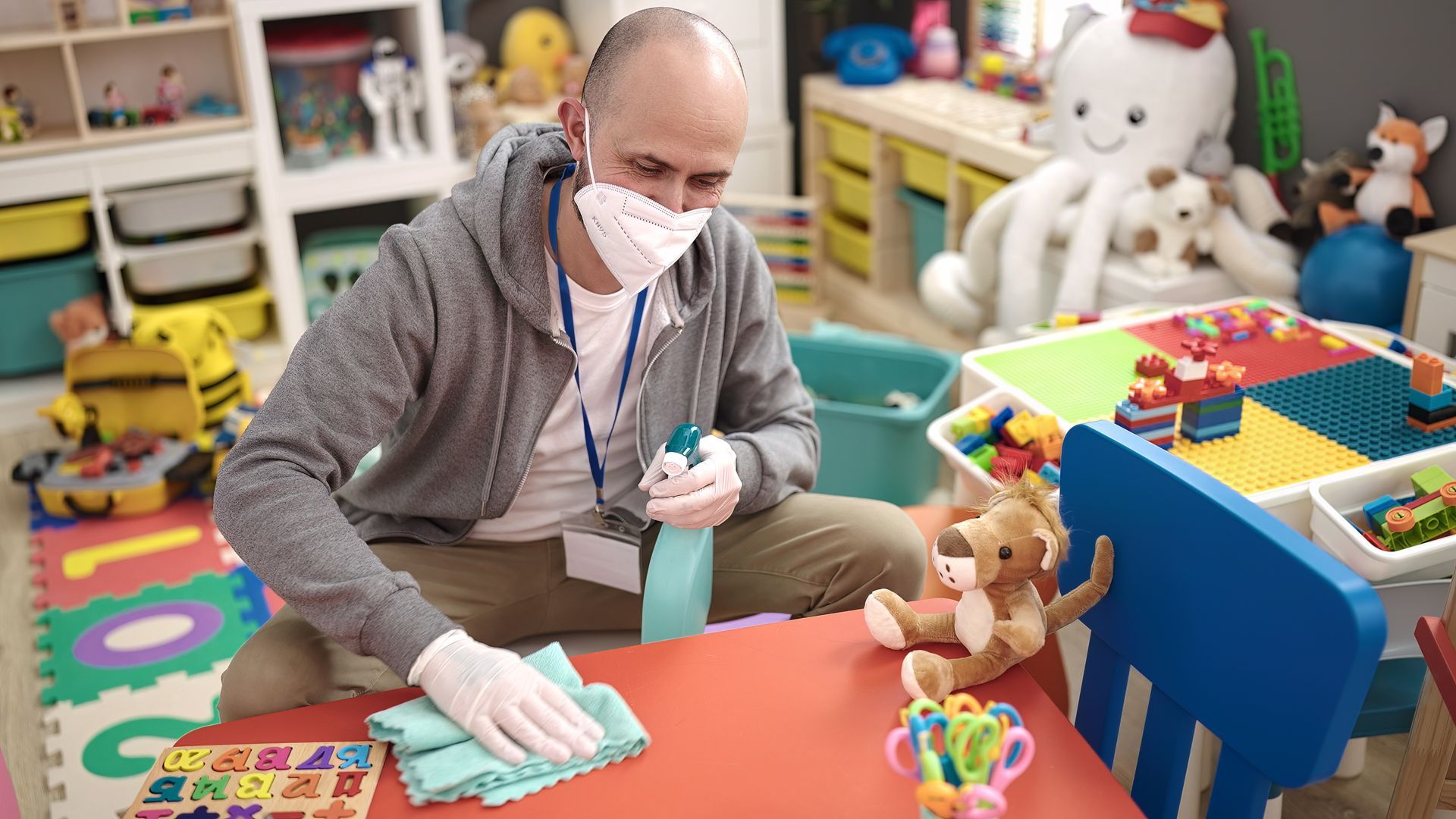 Man in mask and gloves disinfecting a playroom table with spray bottle and cloth.