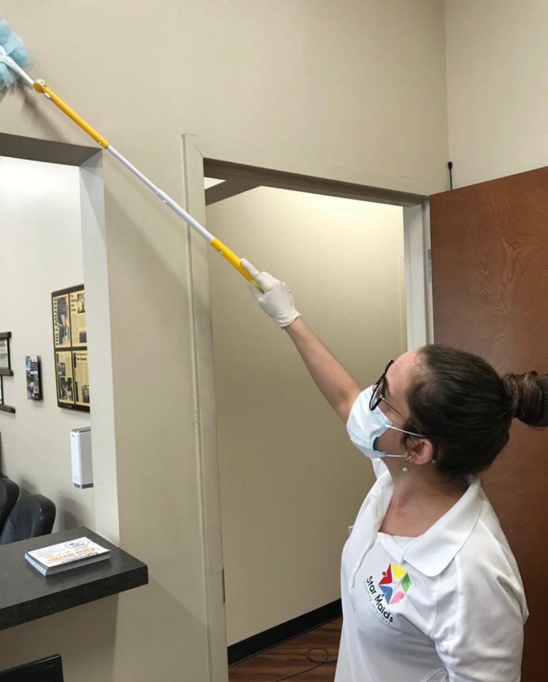 Person dusting a doorway with an extendable duster; wearing gloves, mask, and uniform; cleaning an office. Person dusting a doorway with an extendable duster; wearing gloves, mask, and uniform; cleaning an office.