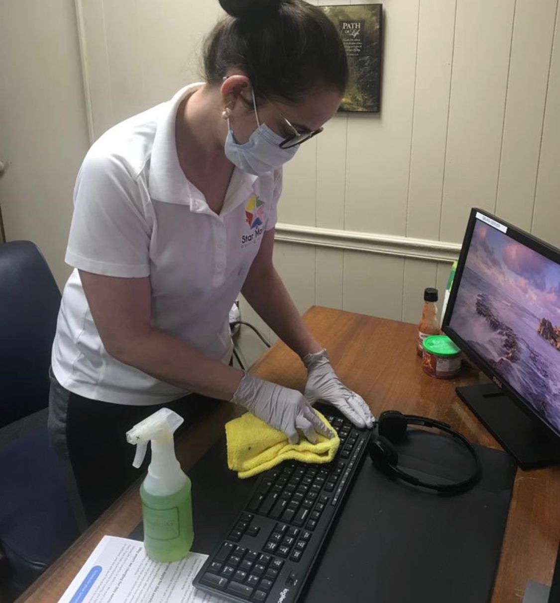 Person wearing mask and gloves cleaning a keyboard with a spray bottle and cloth on a desk. Person wearing mask and gloves cleaning a keyboard with a spray bottle and cloth on a desk.