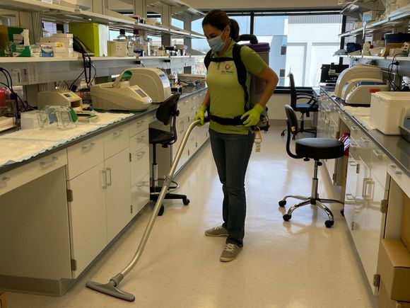Person wearing a mask vacuuming a lab floor with a backpack vacuum cleaner.