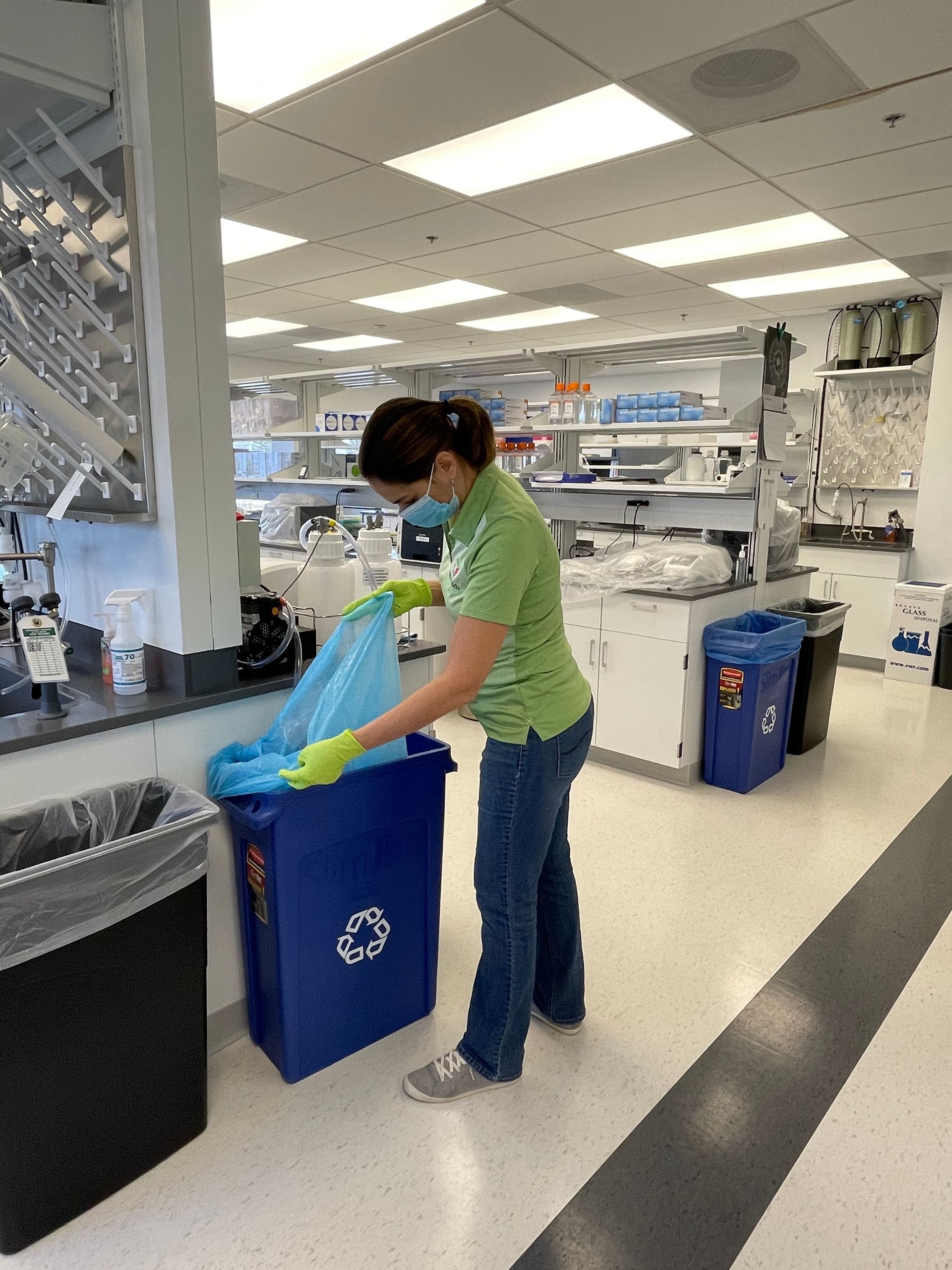 Person in a lab emptying a blue recycling bin into a larger bag. The person wears a mask and gloves.