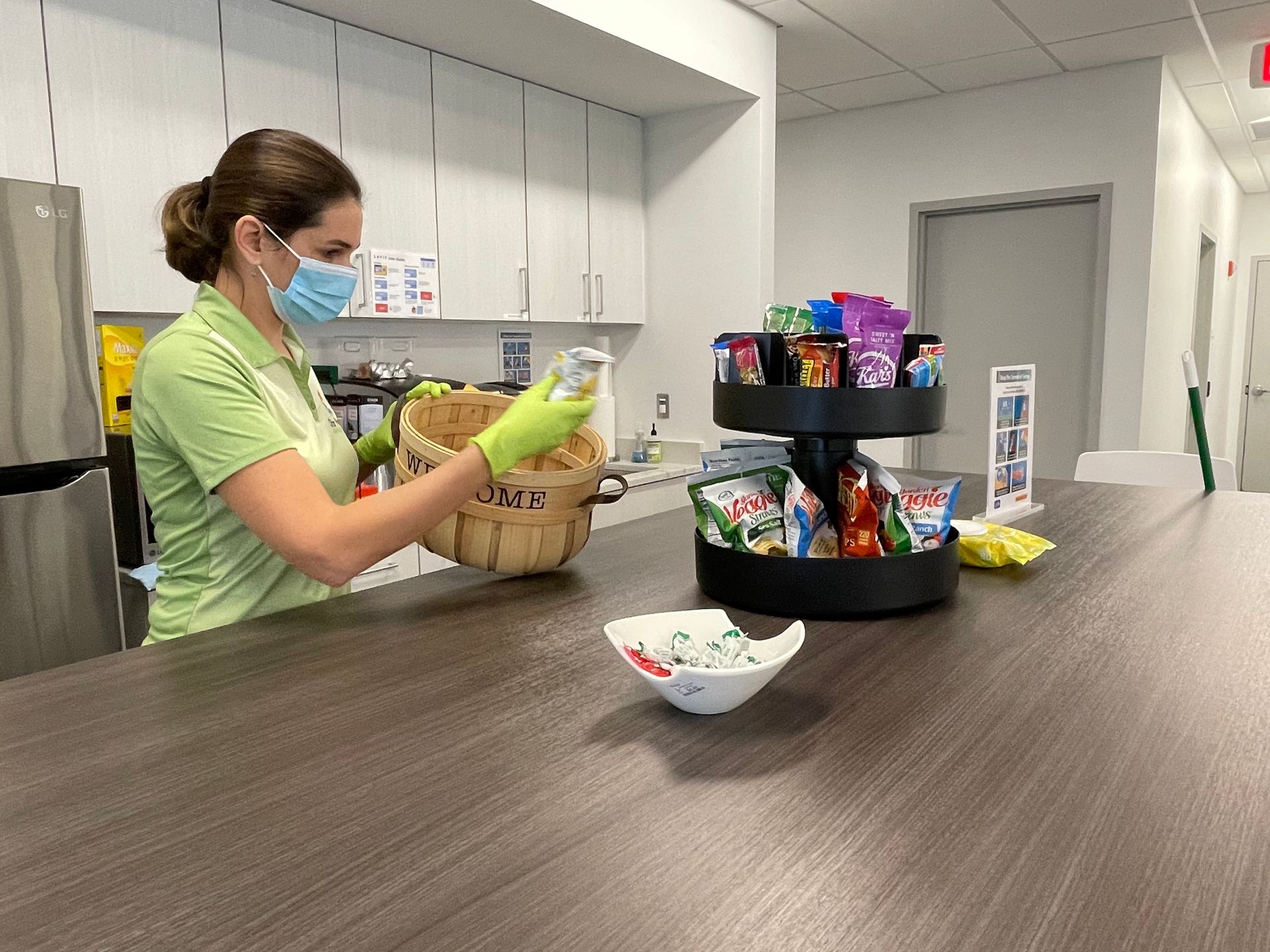 Woman in mask and gloves putting items from a basket into a jar next to a snack display on a countertop. Woman in mask and gloves putting items from a basket into a jar next to a snack display on a countertop.