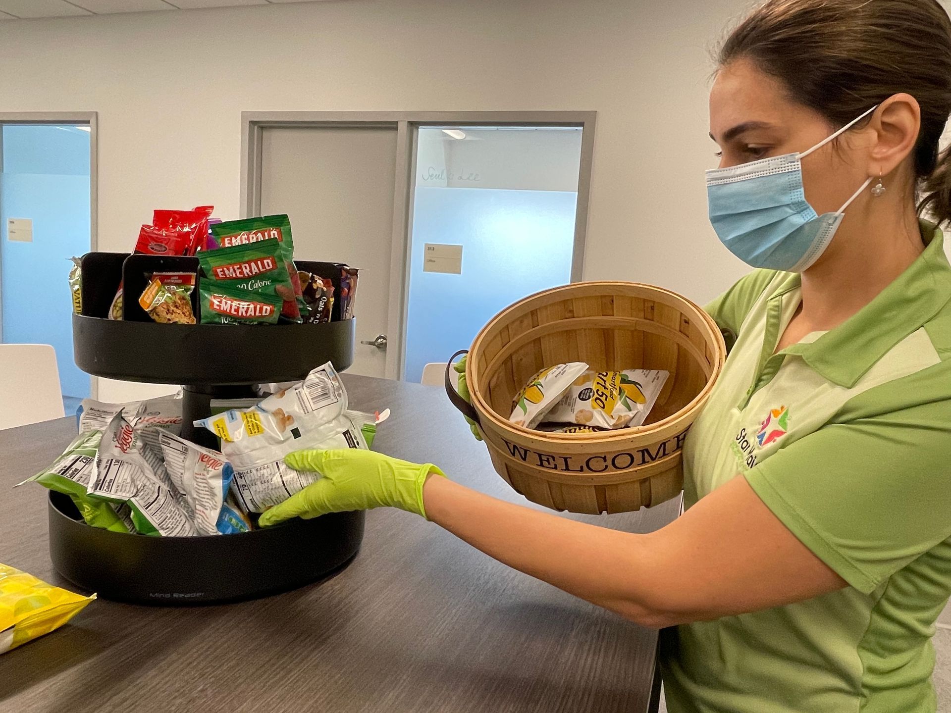 Woman in mask and gloves arranges snacks on a two-tiered carousel and in a basket in an office setting. Woman in mask and gloves arranges snacks on a two-tiered carousel and in a basket in an office setting.