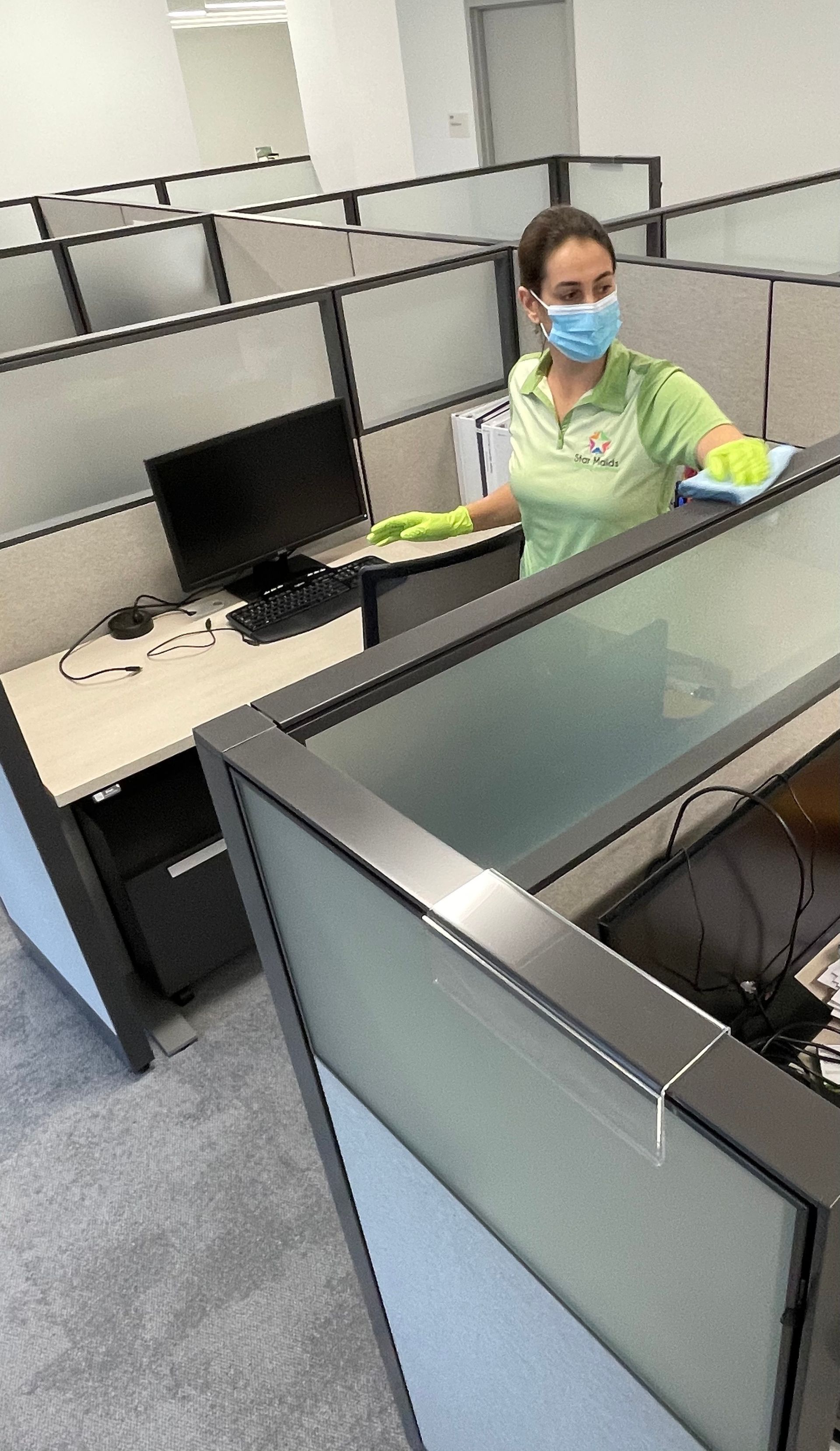 Person wearing mask and gloves cleaning office cubicle; gray walls and carpet.