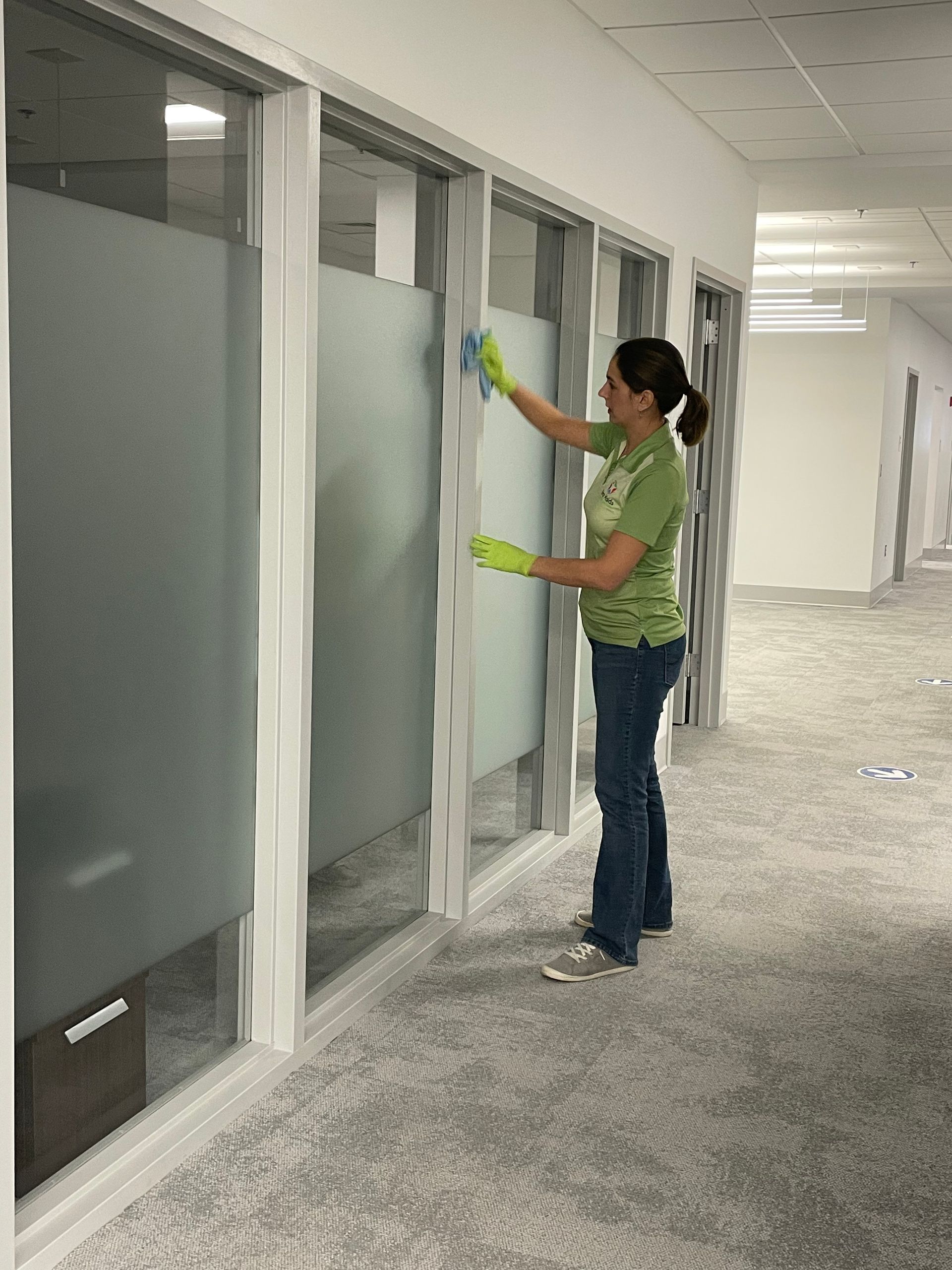 Woman in green shirt, jeans, and gloves cleaning a glass office partition in a hallway with a gray carpet.