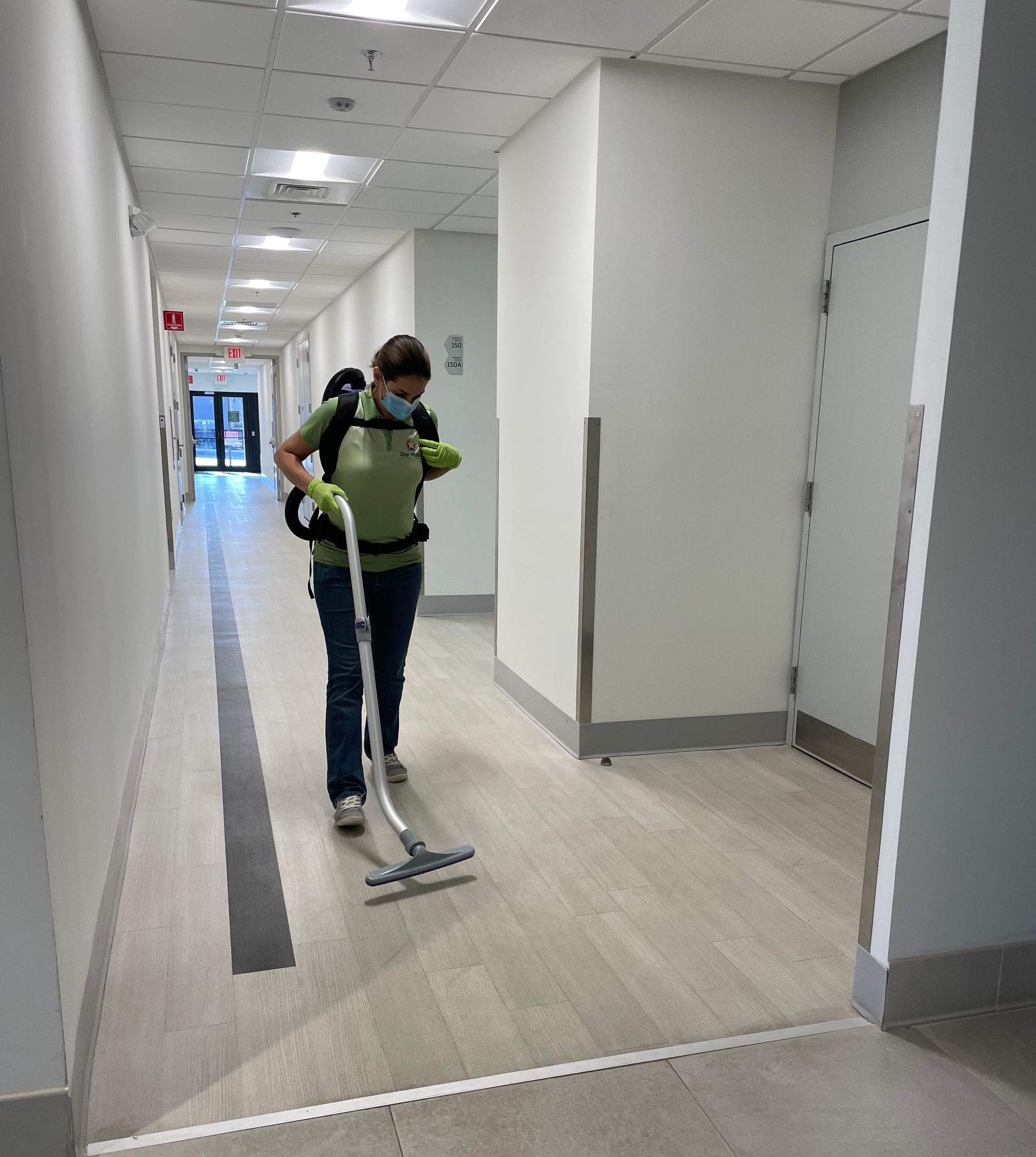 Person vacuuming a long hallway. The person wears a mask and a backpack vacuum. Person vacuuming a long hallway. The person wears a mask and a backpack vacuum.