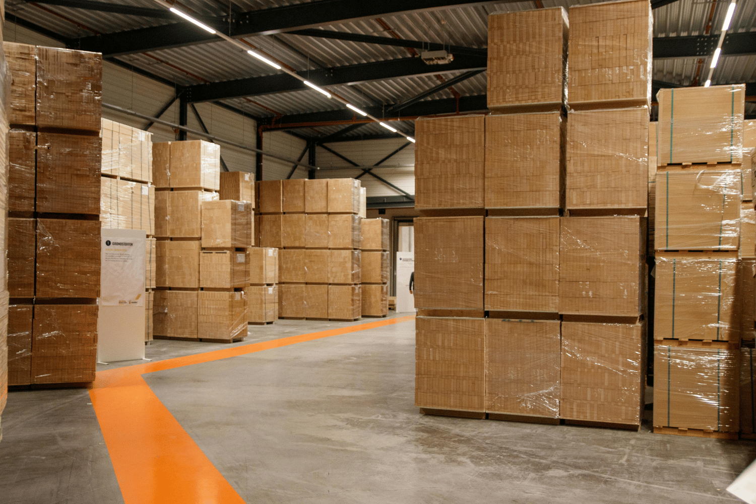 Warehouse interior with stacks of packaged goods, an orange path, and industrial lighting.