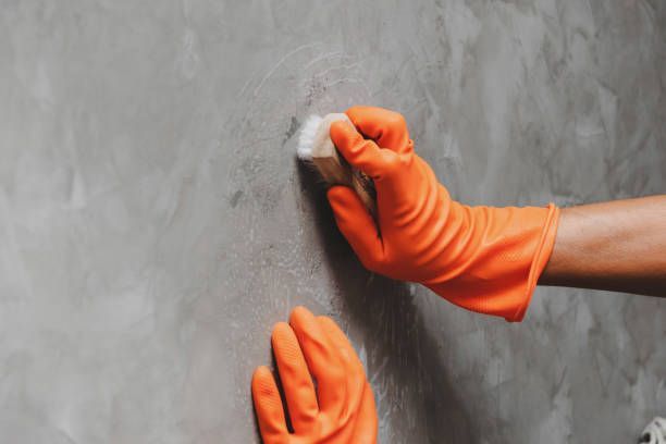 A pair of hands wearing orange gloves clean a concrete or stone surface with a scrubbing brush.