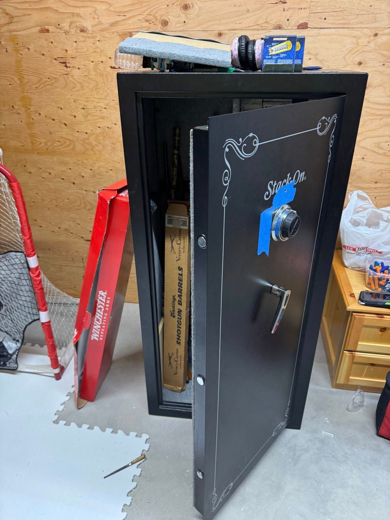 Black gun safe with open door; contains rifles. Set in a cluttered room.
