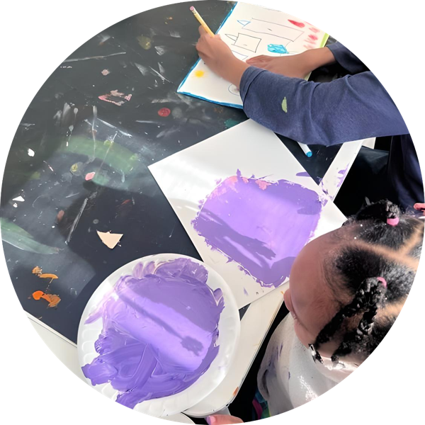 A little girl is sitting at a table painting with purple paint