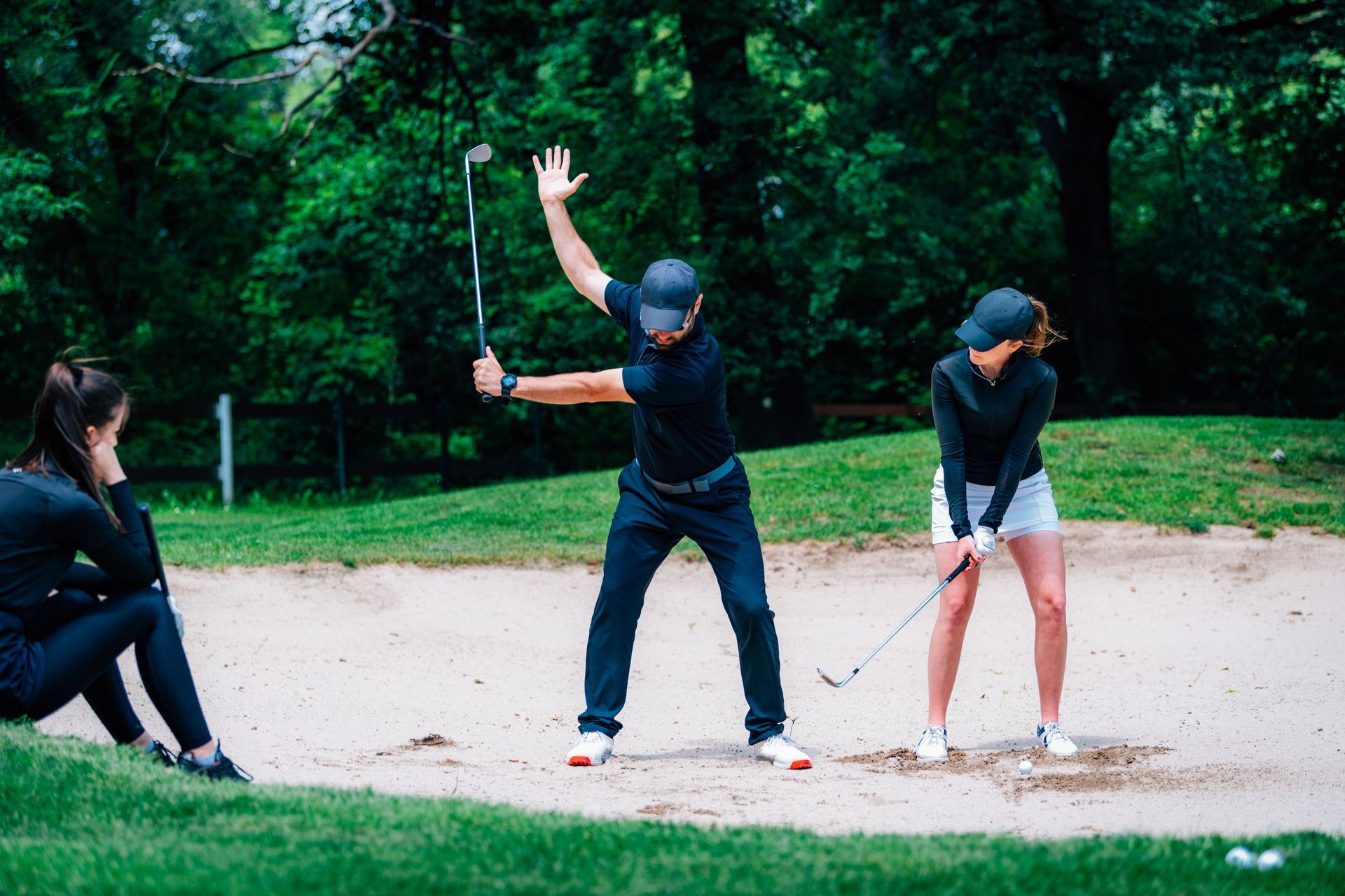 Man demonstrating a golf swing to woman in a sand trap, another woman watches.