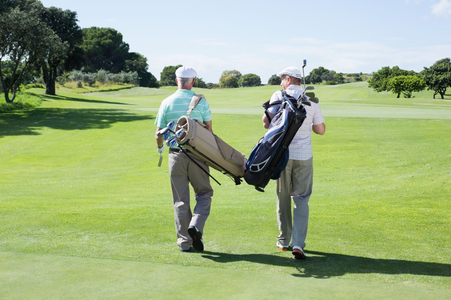Two men in golf attire walking on a green fairway, carrying golf bags under a sunny sky.
