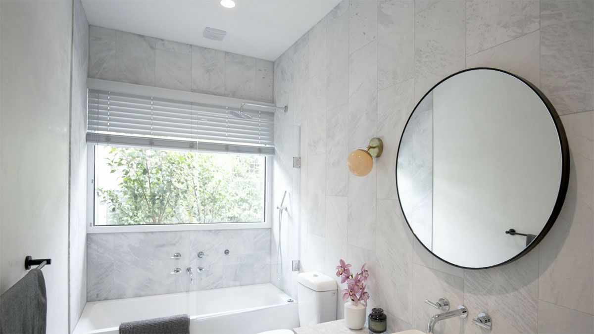 Modern bathroom with marble walls, round mirror, and a window with a view of greenery.