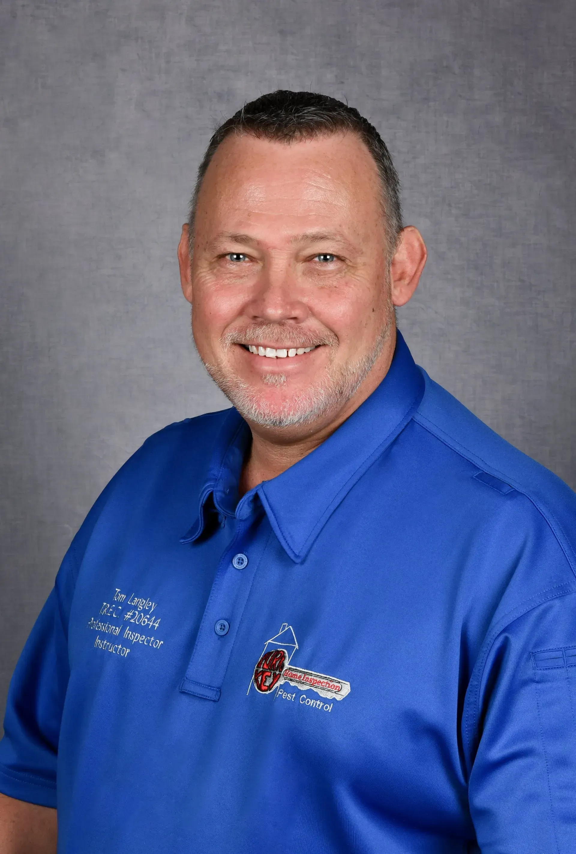 A person smiling, wearing a blue collared shirt with an embroidered logo, set against a grey background.