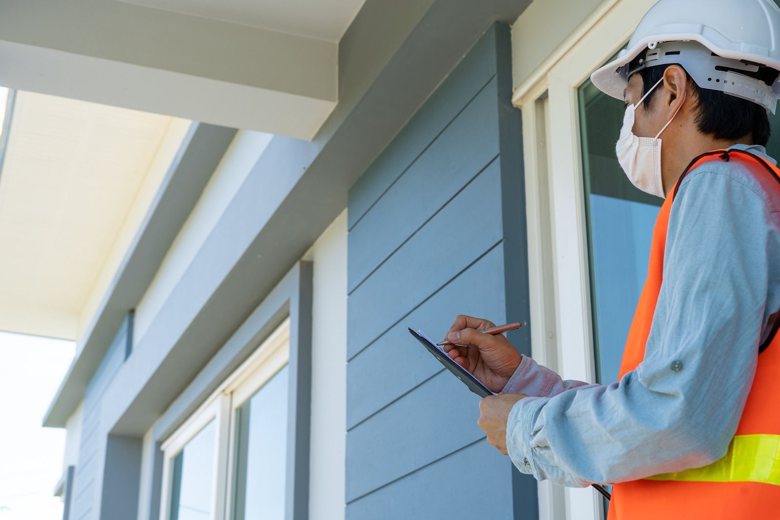 A person in a hard hat, face mask, and high-visibility vest inspects a building exterior while writing on a tablet.