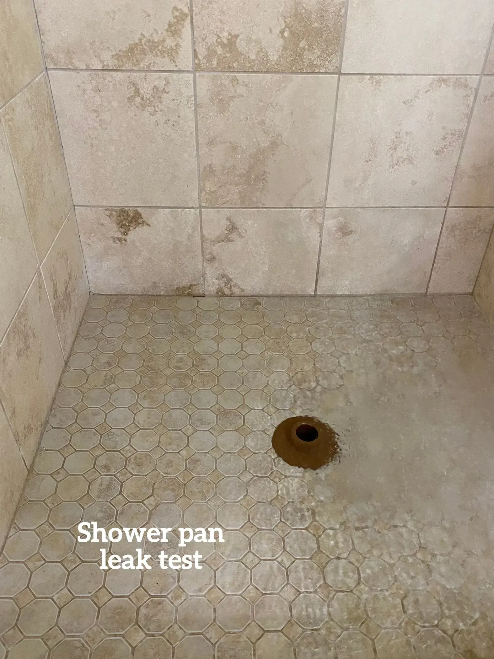 A shower stall floor undergoing a leak test, with water pooled around a central drain and beige tiled walls.
