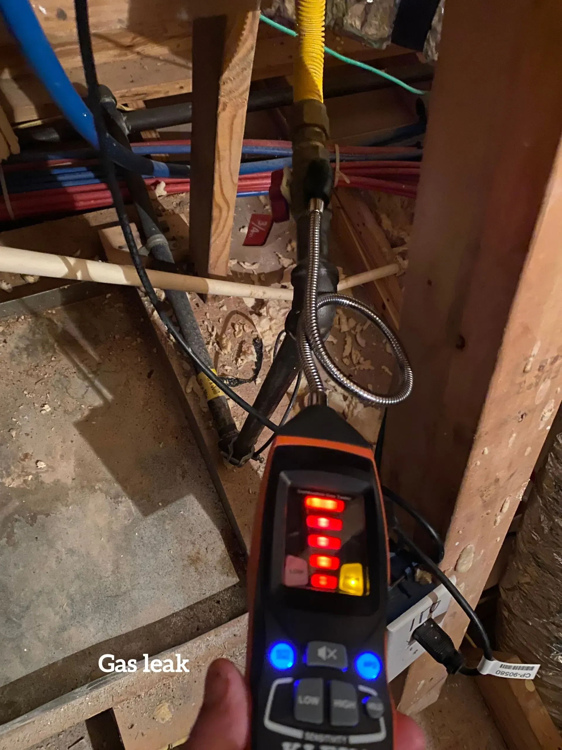 A handheld combustible gas detector with red lights activated near a metal gas pipe connection in an unfinished space.