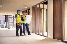 Two construction workers in high-visibility vests and hard hats review architectural plans inside an unfinished building.