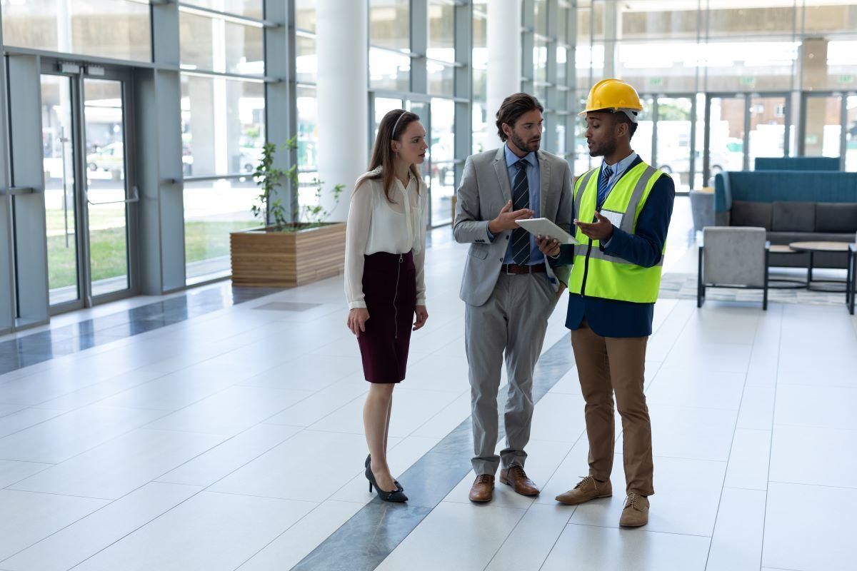 A business professional and a worker in a high-visibility vest and hard hat discuss a document in a modern building lobby.