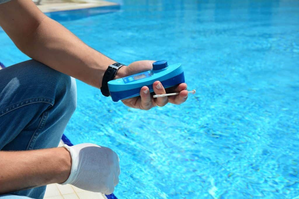 A person in jeans and a glove tests swimming pool water with a blue digital photometer beside a clear blue pool.