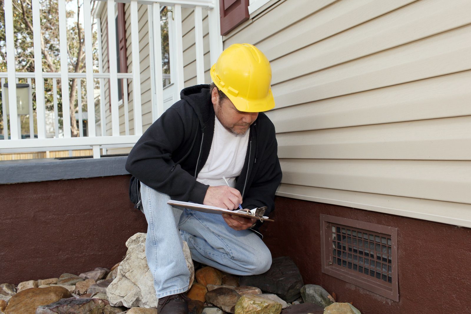 A construction professional in a hard hat kneeling by a house foundation, writing notes on a clipboard.