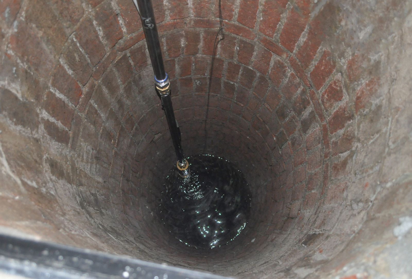 A deep, cylindrical brick well with a black pipe extending into dark water at the bottom.