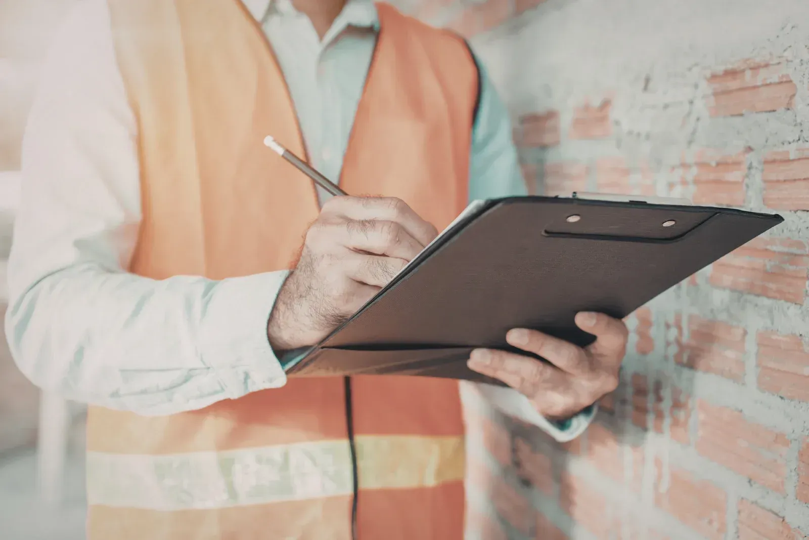 A person in an orange safety vest writing on a clipboard against an unfinished brick wall.