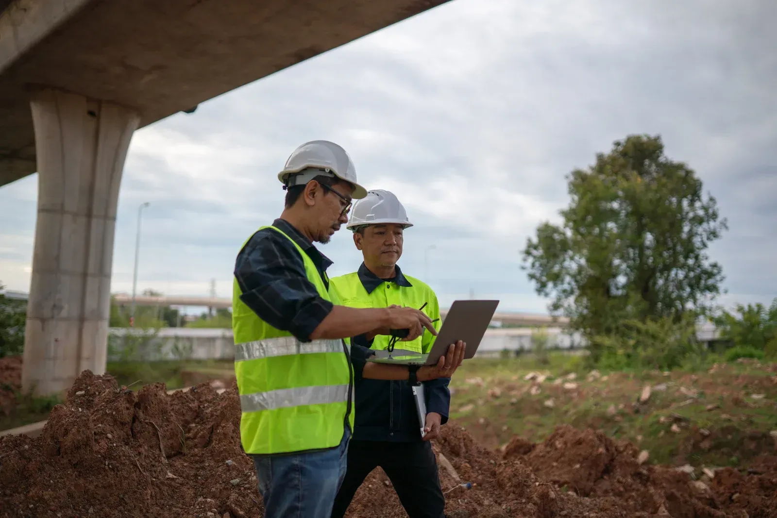 Two construction professionals in hard hats and high-visibility vests review data on a laptop at a construction site.