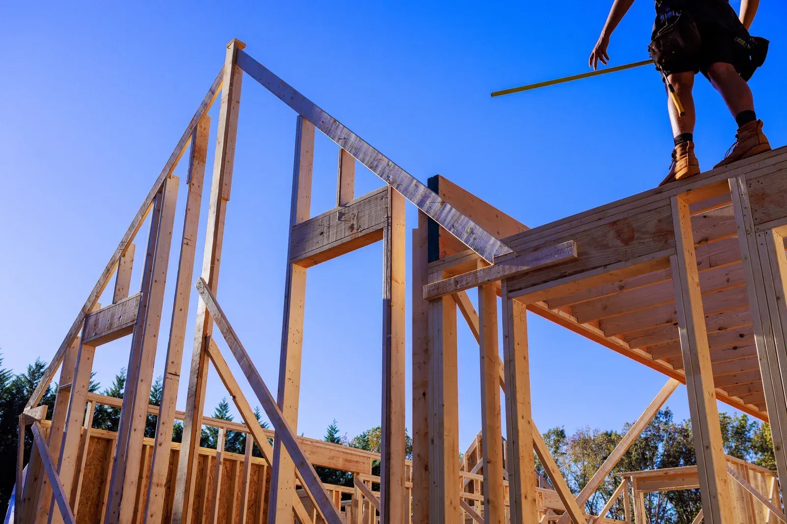 A low-angle view of a wooden house frame under a bright blue sky, with a worker standing on the upper level.
