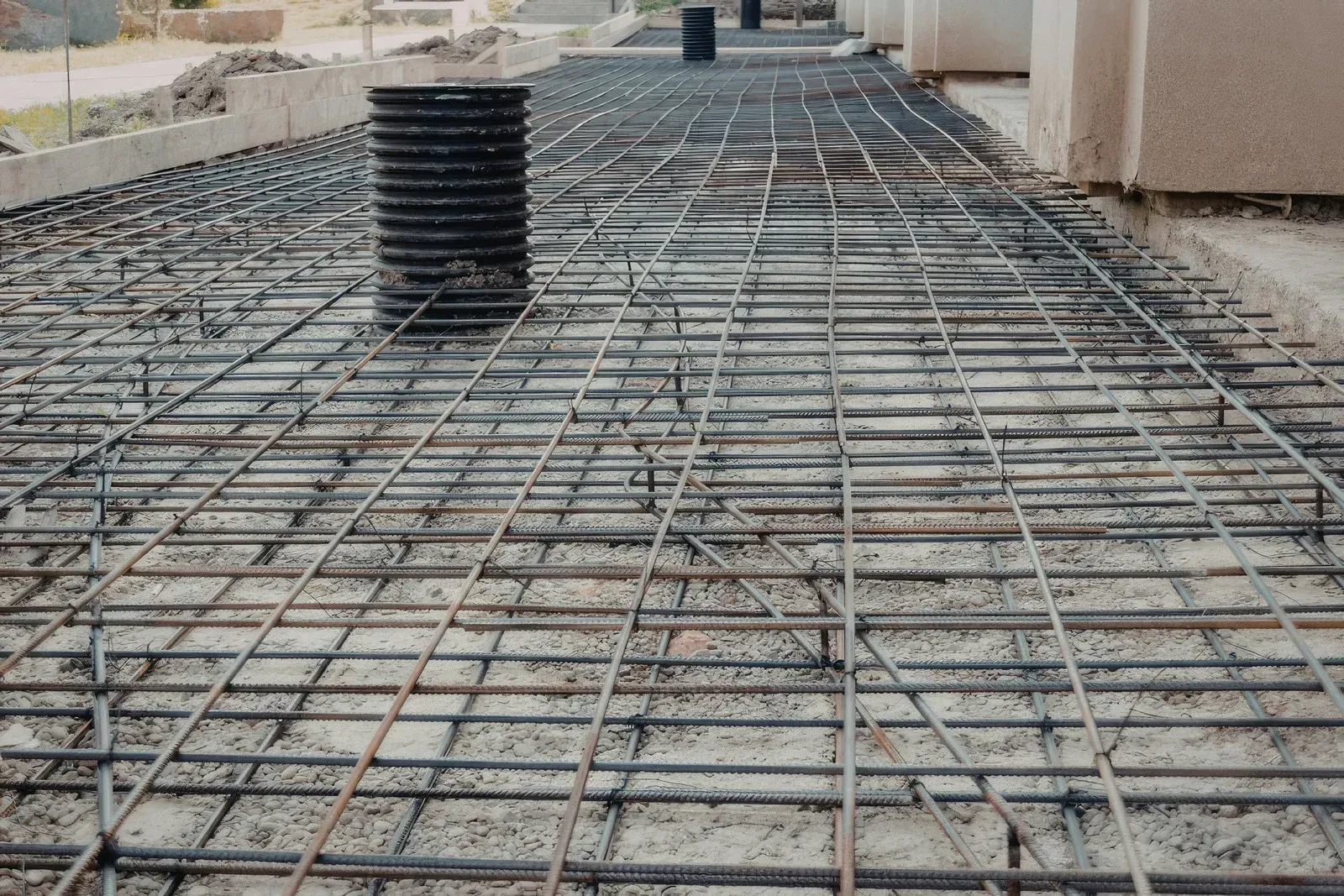 Rebar grid laid out on a gravel construction site, prepared for a concrete pour near a building foundation.