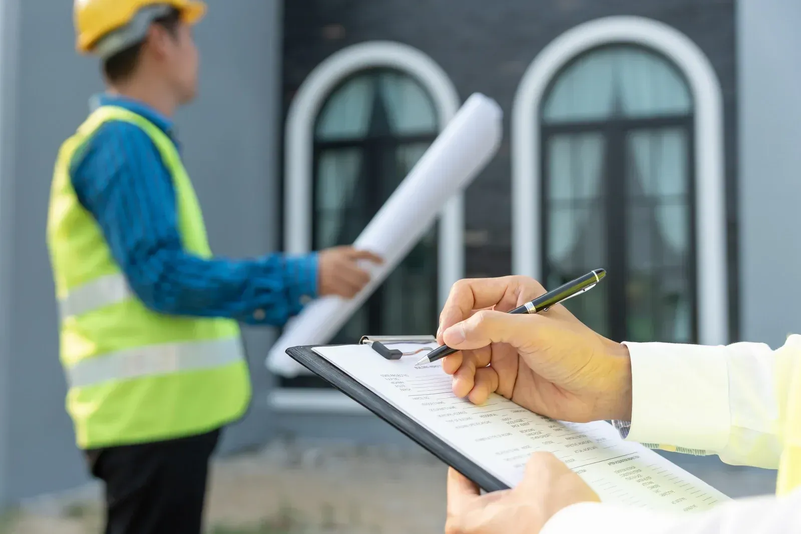 A person holding a pen writes on a clipboard in the foreground while another person in a safety vest holds blueprints.