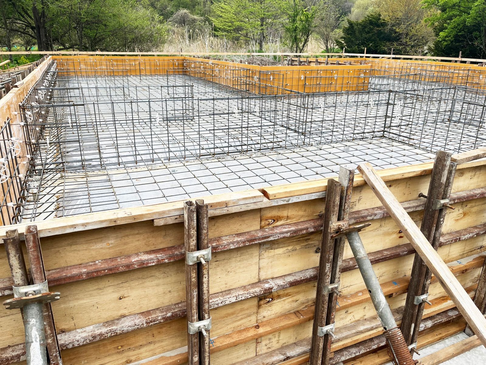 A construction site shows a reinforced concrete foundation, with steel rebar grids inside wooden formwork.
