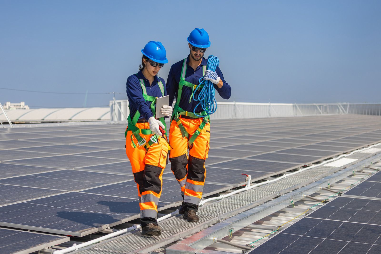 Two technicians in safety gear and blue hard hats walk across a rooftop covered in solar panels while holding equipment.