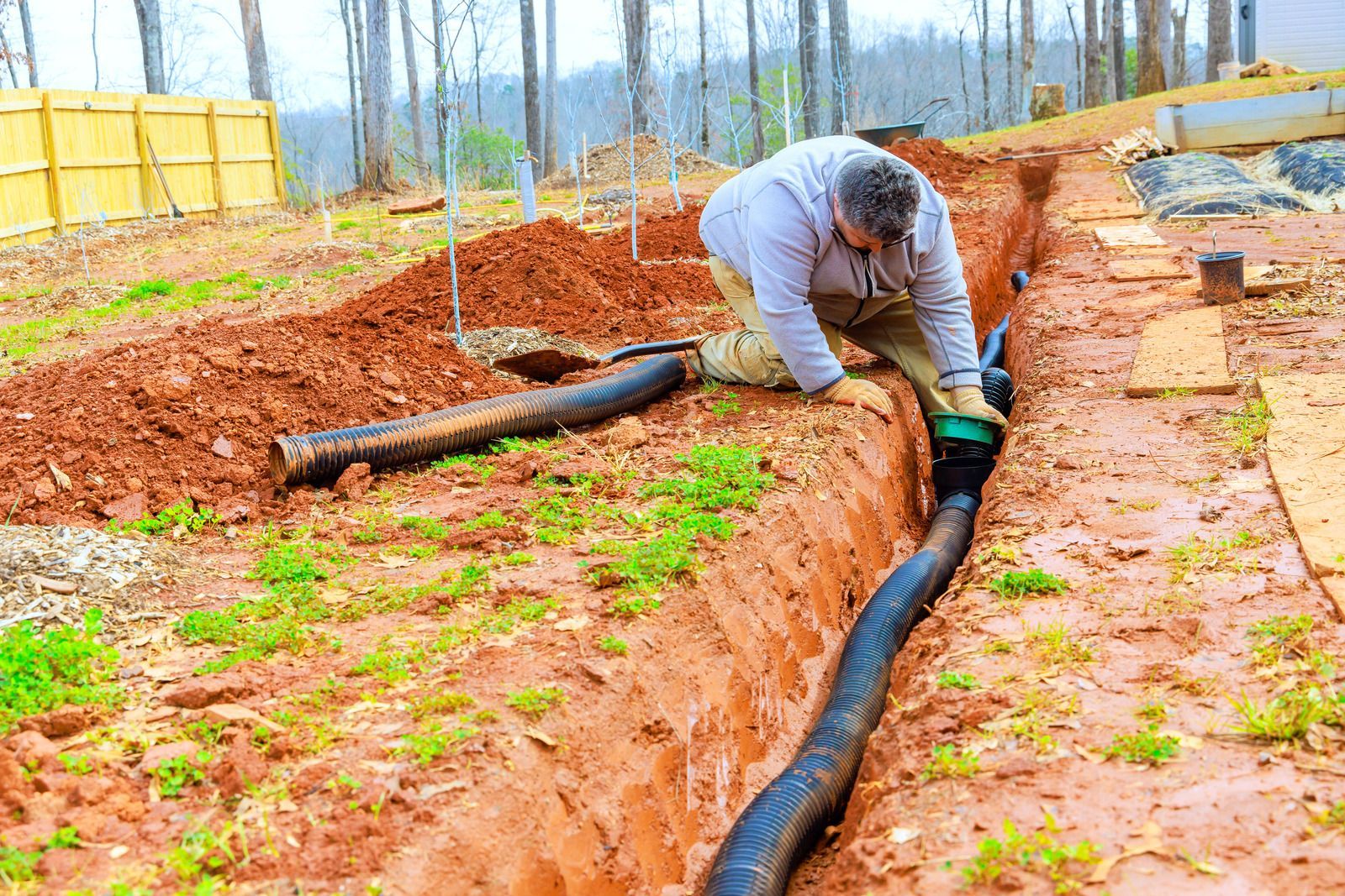 A person in a gray jacket kneels in a red-dirt trench, working on a corrugated black drainage pipe at a construction site.